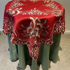 May include: Red table cover with gold and white embroidered floral designs. The cover has a scalloped edge and is draped over a green table.