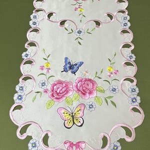 Puede incluir: Camino de mesa blanco bordado con borde festoneado. Presenta rosas rosas, mariposas azules y amarillas y detalles florales. El camino de mesa tiene un diseño de lazo rosa y está decorado con flores azules y rosas.