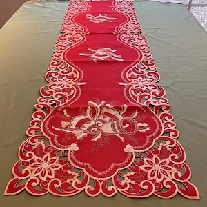 Puede incluir: Caminero de mesa rojo con un diseño bordado en oro de campanas y acebo. El camino de mesa tiene un borde festoneado con un delicado patrón de encaje.