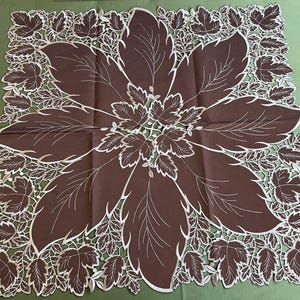 May include: A brown square tablecloth featuring a large floral design. The central design is surrounded by a white leaf pattern. The tablecloth is made of a soft material, possibly linen.