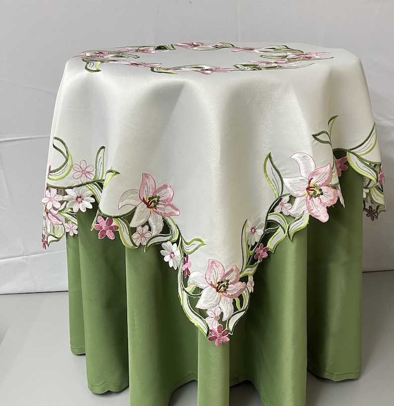 May include: White table cloth with a green and pink floral embroidery design. The tablecloth is laid on a green table.
