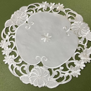 以下が含まれることがあります： 白く円形のドイリーで、複雑な花のレース模様が施されています。中央には小さな花があり、縁には大きく詳細な花の模様が飾られています。ドイリーは緑色の背景に配置されています。