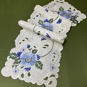 May include: White embroidered table runner with a floral design. The runner features large blue flowers with green leaves and smaller blue flower accents. The edges are scalloped and the design is intricate.