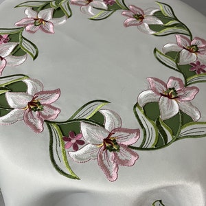 May include: White table runner with a green and pink embroidered floral design. The design features a repeating pattern of lilies with green leaves.