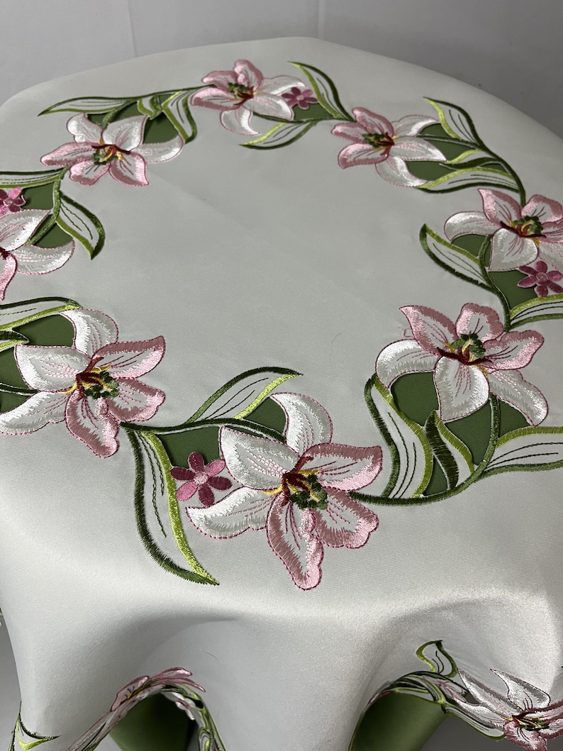 May include: White table runner with a green and pink embroidered floral design. The design features a repeating pattern of lilies with green leaves.