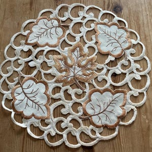 May include: Circular cream-coloured doily with an openwork pattern and embroidered leaves in cream and gold. The leaves feature brown stitching, creating a decorative accent for home decor.