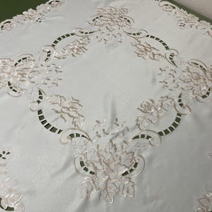 May include: White tablecloth with delicate embroidered floral designs in a cream color. The tablecloth has a scalloped edge and a square shape.