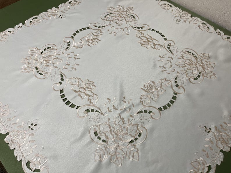 May include: White tablecloth with delicate embroidered floral designs in a cream color. The tablecloth has a scalloped edge and a square shape.