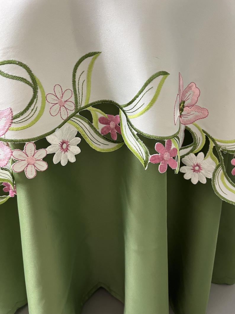 May include: White tablecloth with green and pink embroidered floral design. The tablecloth has a green scalloped edge.