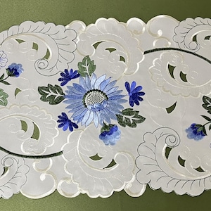 May include: White embroidered table runner with a delicate floral design featuring blue and green flowers. The runner has a scalloped edge and a cut-out pattern.