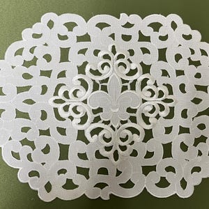 May include: White lace table runner with a delicate floral design and a fleur-de-lis in the center.