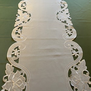 May include: White embroidered table runner with a floral design. The runner has a scalloped edge and is made of a delicate fabric.
