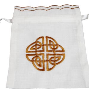 May include: A white drawstring bag with a gold Celtic knot design embroidered on the front. The top edge has a scalloped trim in gold, and white satin ribbons serve as the drawstring.