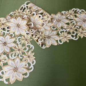 Può includere: Un runner da tavolo ricamato bianco e oro con un motivo floreale. Il runner presenta un delicato motivo a pizzo ed è perfetto per aggiungere un tocco di eleganza a qualsiasi tavola.