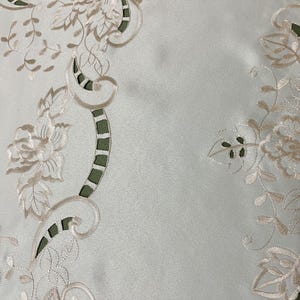May include: White table runner with a delicate floral embroidery design in a cream color. The embroidery features intricate floral patterns, including roses and leaves, along the edges of the runner.
