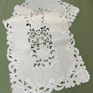 May include: Ivory-colored, embroidered table runner with a floral design. The runner features a delicate, openwork pattern with flowers and scrolling details along the edges and center. The fabric appears sheer and lightweight.