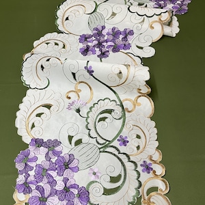 May include: White table runner with an intricate floral design featuring purple and green embroidered flowers. The runner has a scalloped edge and is perfect for adding a touch of elegance to any table setting.