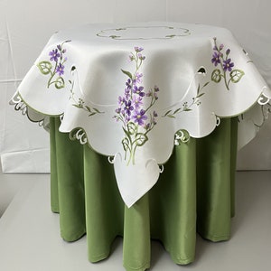 Può includere: Tovaglia bianca con un ricamo floreale con fiori viola e verdi. La tovaglia ha un bordo a festoni ed è drappeggiata su un tavolo verde.