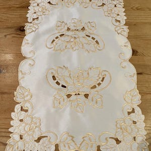 May include: White table runner with gold embroidered floral design. The design features intricate leaves and flowers.
