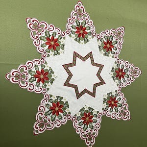 Könnte beinhalten: Ein weiß-roter Tischläufer in Poinsettienform mit sternförmiger Mitte. Der Läufer zeigt gestickte rote Poinsettien, grüne Blätter und dekorative Schnörkel. Der Läufer liegt auf einer grünen Oberfläche.