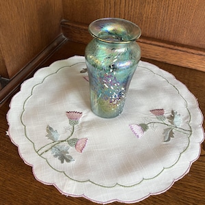 May include: A decorative iridescent glass vase with a flared rim sits on a white, scalloped-edge doily. The doily features embroidered thistle flowers in pink, green, and gray. The vase has a textured surface and a gold rim.