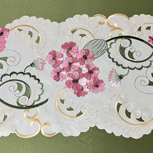 May include: White table runner with a delicate floral design. The runner features pink flowers and green leaves on a white background with gold accents.