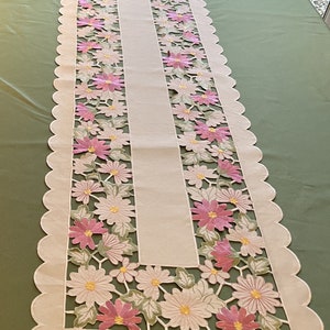 Puede incluir: Un camino de mesa blanco con un borde festoneado y flores bordadas en rosa y verde. El camino de mesa tiene un diseño recortado, revelando la superficie de la mesa debajo.