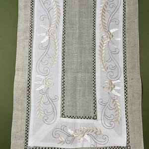 Può includere: Un runner da tavolo in lino bianco con ricami floreali in beige e bianco. Il runner ha un delicato bordo in pizzo.