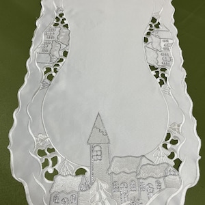May include: White table runner with a silver embroidered winter village scene. The design features a church, houses, trees, and snowflakes.