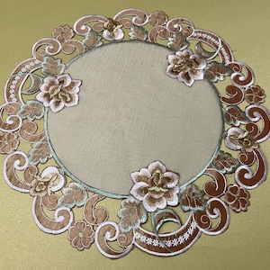 May include: Round, embroidered placemat with a floral design. The placemat features a central beige circle surrounded by a border of brown and white embroidered flowers and scrollwork. The placemat is approximately 12 inches in diameter.