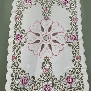 May include: A rectangular white table runner featuring intricate cutwork lace borders, embroidered pink rose motifs, and a large central floral design outlined in pink thread. The edges are scalloped, and the fabric has a smooth, satin-like finish.