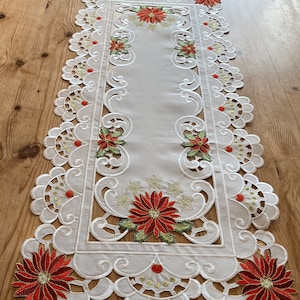May include: White table runner with a delicate lace design and red poinsettia embroidery. The runner features a scalloped edge and a central panel with a repeating floral pattern.