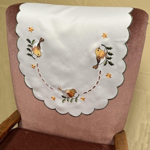 以下が含まれることがあります： 白い椅子の背もたれカバー。スカラップエッジで、緑の枝にとまった3羽の茶色と黄色の鳥が刺繍されています。カバーは小さな黄色の花と点線で飾られています。