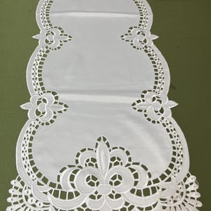 Puede incluir: Camino de mesa blanco con intrincados patrones florales y geométricos calados. El camino de mesa presenta un panel central con bordes decorativos y detalles florales. El diseño es simétrico, con patrones calados detallados en los lados y extremos.