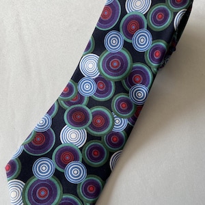Puede incluir: Una corbata con fondo negro y un patrón repetitivo de círculos concéntricos en tonos morado, verde, azul claro, blanco y rojo. Los círculos varían en tamaño y se superponen a lo largo de toda la superficie de la tela.