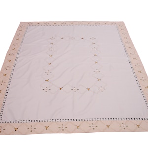 May include: A rectangular white tablecloth with a beige border. The border features a repeating pattern of small flowers and triangles. The center of the tablecloth has a floral design. The tablecloth is made of a soft fabric.