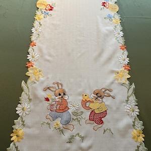 Può includere: Runner da tavola pasquale color crema con ricami di coniglietti, fiori e uova di Pasqua. Il runner presenta un bordo smerlato con accenti floreali gialli, bianchi e arancioni. I coniglietti portano fiori e un pulcino.