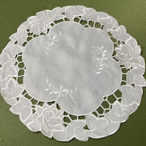 May include: A white, round doily with a floral and leaf design. The center is a solid white, surrounded by a scalloped edge with embroidered flowers and leaves. The doily is approximately 10 inches in diameter.