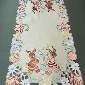 May include: White embroidered table runner with a green, white and yellow floral border. The centre features two brown bunnies holding pink and red Easter eggs. The border also features red and orange Easter eggs.