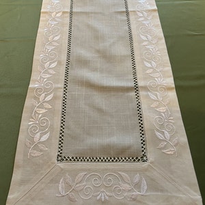 May include: A white linen table runner with an embroidered floral design along the edges. The design features white leaves and swirls.