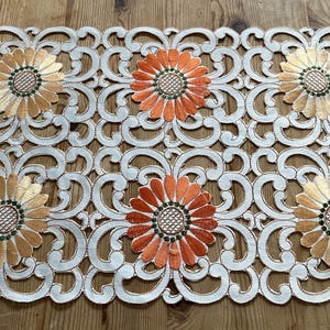 May include: White embroidered table runner with a delicate floral pattern. The flowers are orange, gold, and green. The runner is perfect for adding a touch of elegance to any table setting.