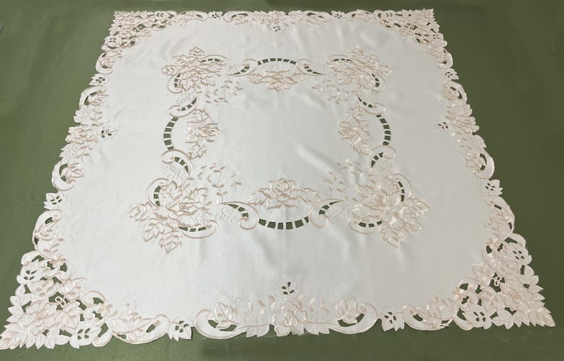 May include: White embroidered tablecloth with a floral design. The tablecloth has a delicate lace pattern around the edges and features a central floral motif.