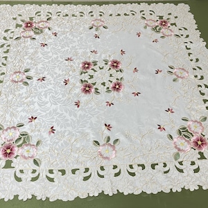 Könnte beinhalten: Weiße Tischdecke mit einem zarten floralen Stickmuster. Die Tischdecke hat einen gewellten Rand und komplizierte Blumenmuster in Rosa- und Grüntönen.