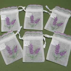 May include: Six white organza sachets with embroidered lavender sprigs. Each sachet has a drawstring closure and a scalloped edge with green and purple trim. The lavender design is purple with green stems.