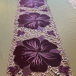 May include: A purple table runner with a delicate lace design and three embroidered hibiscus flowers. The runner is perfect for adding a touch of elegance to any table setting.