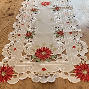 May include: White table runner with a delicate lace design and red poinsettia embroidery. The runner features a scalloped edge and a central design of poinsettias with green leaves.