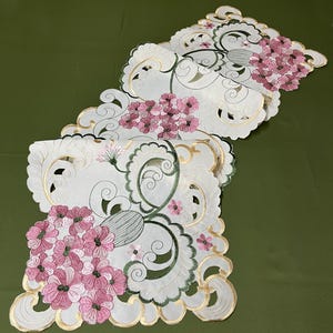 May include: White table runner with a delicate floral design. The runner features pink flowers and green leaves embroidered on a white background. The edges of the runner are scalloped and have a gold trim.