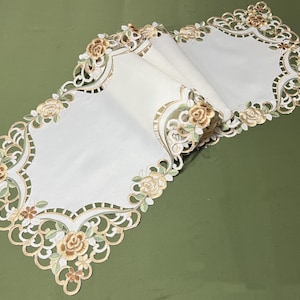 May include: Ivory-colored table runner with a floral design. The runner features embroidered roses in shades of beige and green, with a scalloped edge. The runner is folded on a green surface.