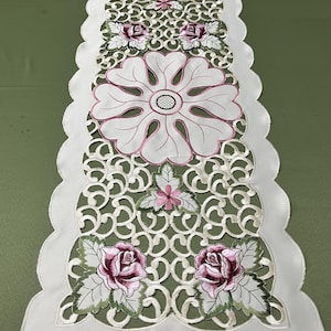 May include: White table runner with embroidered floral and scrollwork design. Features large white flowers with pink outlines and centers, and pink and green rose accents. The edges are scalloped.
