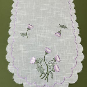 Peut inclure: Un chemin de table rectangulaire blanc à tissage texturé, doté de bords festonnés soulignés par un fil violet et vert. Le chemin de table est orné de fleurs de chardon violettes brodées et de feuilles vertes disposées en bouquet.